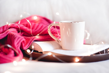 Cup of tea staying on open book with knitted sweater on wooden tray in bed closeup. Good morning. Breakfast.