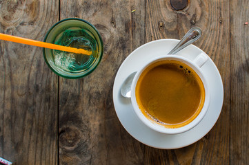 Strong and dark coffee in a white porcelain cup served with a glass of water on a side on a wooden table outdoors