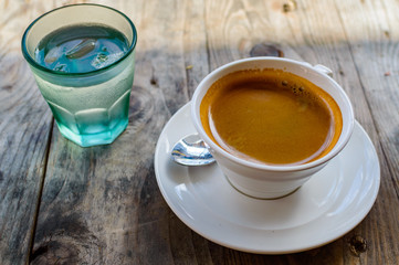 Strong and dark coffee in a white porcelain cup served with a glass of water on a side on a wooden table outdoors