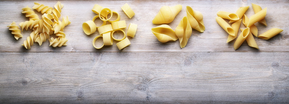 Raw pasta: fusilli, paccheri, conchiglie, and penne on wooden background. Top view, space for text.