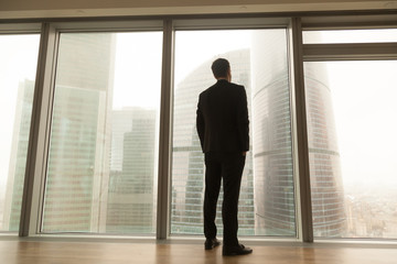 Contemplative businessman standing near big window looking out at city, thoughtful entrepreneur building future plans, thinking over ways to overcome business problems, pass through crisis, back view