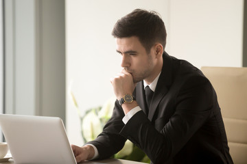 Serious focused businessman working on laptop computer online, looking at screen, trader monitoring real time cryptocurrency exchange rates, using business management software, browsing web services