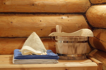 Fototapeta na wymiar Bath accessories in the interior of the Russian bath. Towels, hat and a wooden bucket and ladle on a background of a log walls in a Russian bath.