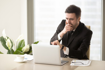 Tired bored businessman yawning at workplace near laptop, looking at wristwatch, checking time,...