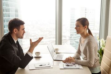 Enthusiastic businessman and businesswoman discussing company business, talking about new project, sharing ideas during meeting sitting at office desk near big window with urban cityscape, side view