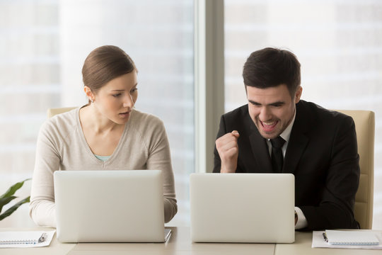 Resentful employee loser looks enviously at promoted colleague winner enjoying success, good news while working on laptop, feels jealous about rivals achievements, team rivalry, unfair competition