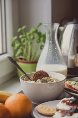 White bowl of cornflakes slice of banana and chocolate cookie on top. Breakfast set in background with milk and coffee