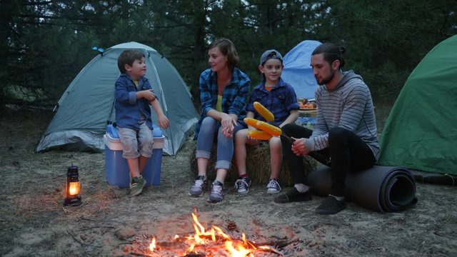 Mother, father and sons on rest in forest, mom, children and dad around fire roasting corn, family evening picnic outdoors, American family during summer holidays, travel in camp