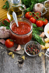 tomato sauce in a glass jar and ingredients on a wooden background, top view, vertical