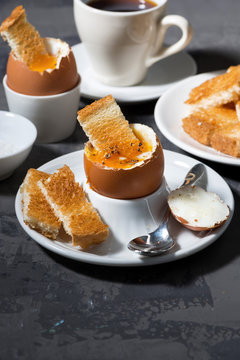 soft boiled egg, toasts and cup of coffee for breakfast on dark background, vertical