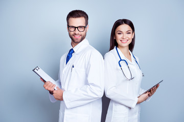 Two medic colleagues in white coats on pure background, holding clipboard and tablet. They are so...