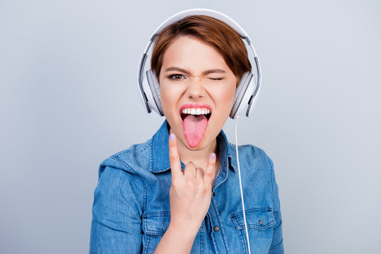 Young woman in jeans shirt in headphones listening to music showing tongue and sign of rock