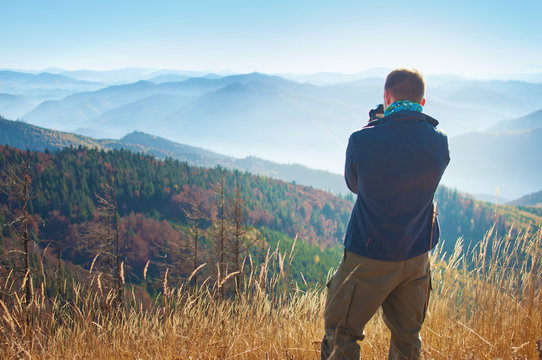 One young man standing with his back turned, holding camera and making photos of a mountain range covered in red, orange, yellow deciduous forest under blue cloudless sky on warm fall day, October