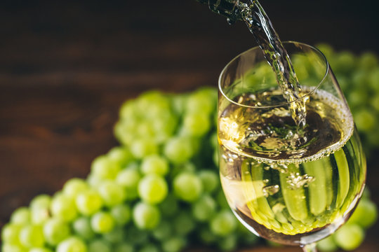 Pouring white wine into a glass with a bunch of green grapes against wooden background