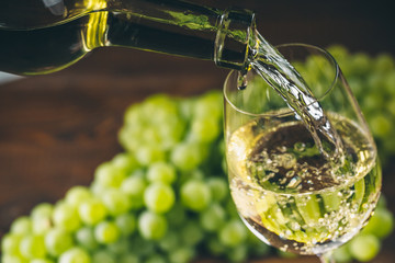 Pouring white wine into a glass with a bunch of green grapes against wooden background