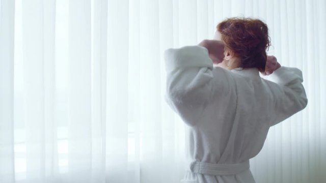 Woman in bathrobe walking to window in hotel room and stretching herself in the morning
