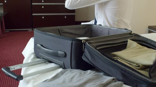 Tilt up of businessman sitting on bed with opened suitcase and clothes in hotel room and talking on the phone after arrival