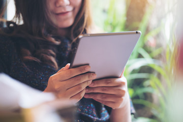A beautiful Asian woman with smiley face holding , using and looking at tablet pc in modern cafe with green nature background
