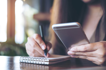 Closeup image of a business woman writing on notebook while using and holding mobile phone on wooden table background in office