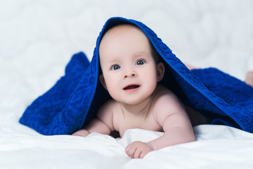 Cute baby girl or boy after shower with towel on head in white sunny bedroom. Child with big blue eyes smiling and relaxing in bed after bath