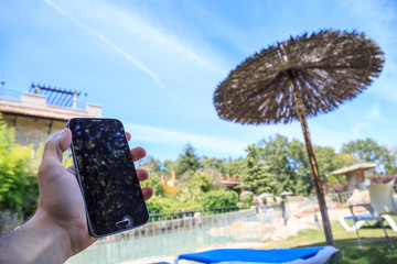 Hand holding a smartphone on a background of a thatched beach umbrella in a pool with a blue sky.   