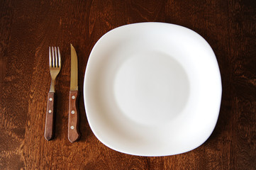 Empty white plate with a fork and knife on a dark wooden brown table