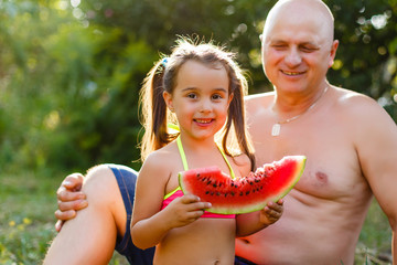 Grandfather and granddaughter eat watermelon and laugh in the garden on the grass in summertime