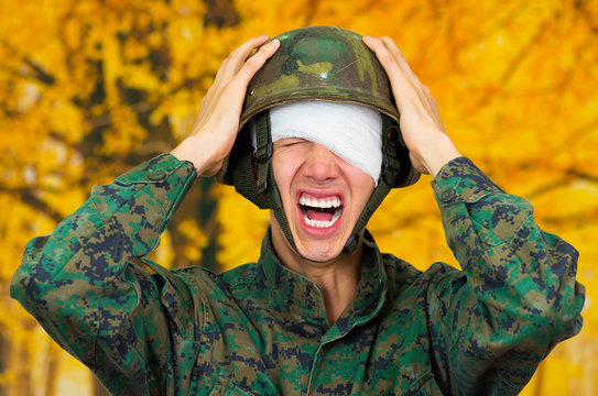 Handsome young soldier wearing uniform suffering from stress, with a white bandage around his head and covering his eye, with both hands touching his helmet and screaming, in a blurred yellow - Powered by Adobe