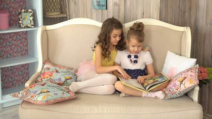 Little girls are reading a book. Two sisters are sitting on the sofa with a book