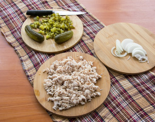 Finely chopped pickled cucumber on the board and a cutting knife; Boiled chicken, onions on the wooden table top on a checkered kitchen towel