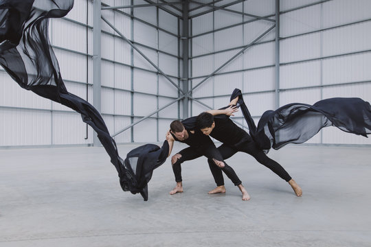 Two male dancers wrestling in a warehouse with black ink-like silk flying around them