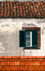 Detail from traditional old Portuguese facade with green and white wooden window, roof with old brown ceramic tiles and old and worn out wall background damaged by exposure to the elements.