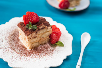 Piece of tiramisu cake with mint and strawberries on a blue background