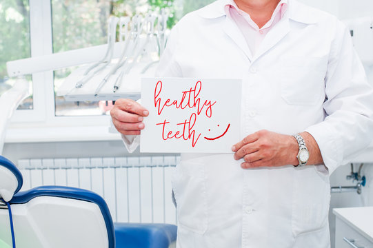 Male Dentist Doctor Holding Blank With The Phrase Heaithy Teeth And Drawn Emoji In His Dental Office. Medical Service And Healthy Life Concept. Space For Text. Selective Focus