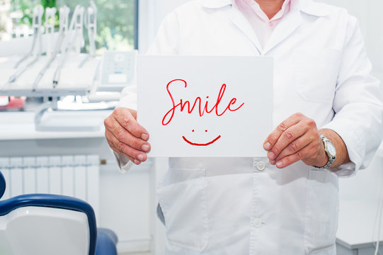 Male Dentist doctor holding blank with word Smile and drawn emoji in his dental office. Medical service and Healthy life concept. Space for text. Selective focus