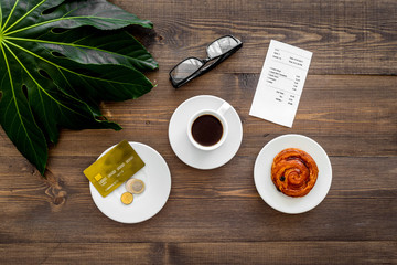 Pay restaurant bill. Bill, bank card, coins near cup of coffee and bun on dark wooden table top view