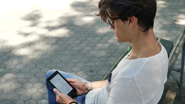 Young attractive man in city park using ebook reader to read a book, sitting on a bench