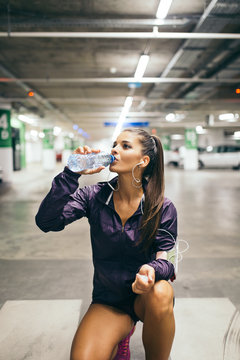 Young woman in public parking garage drinking water after fitness workout..
