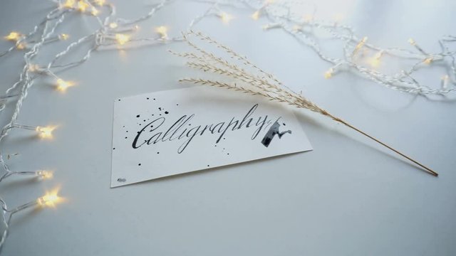 Close-up of Calligraphy inscription in italic fine print on rectangular white sheet of paper, next to table feather grass branch on light table with bright twinkling garland. Concept of fine art and