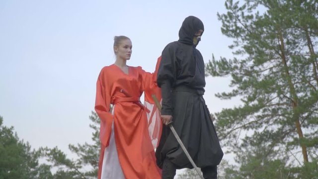 To the ninja in a black kimono from behind comes a geisha in a red dress hugging and picking up katana, slow motion