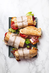 Fresh baguette sandwich bahn-mi styled. Bacon, roasted cheese, tomatoes and lettuce on metallic tray on white marble background. Vertical composition.