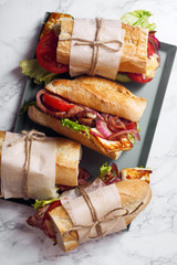 Fresh baguette sandwich bahn-mi styled. Bacon, roasted cheese, tomatoes and lettuce on metallic tray on white marble background.