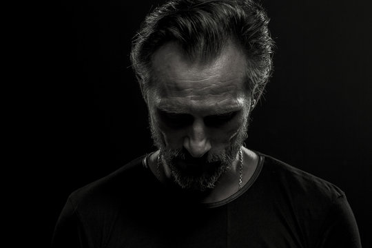 Low key dramatic portrait of mature man on black background. Beardy upset male looking down.