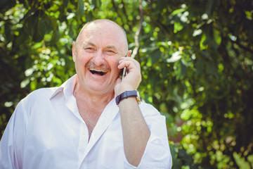 Mature stylish European or American man wear in white shirt at garden, happy senior talk to telephone and hear good news