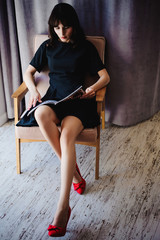 Young attractive woman with long legs in black elegant dress, sits in chair near window in interior of room. Seductive shoes of red light, on beautiful legs