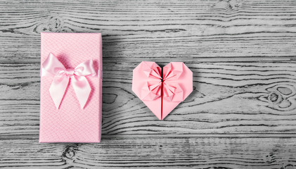 A gift box with a bow and a delicate pink heart made of paper on a gray background.  Postcard heart of origami. Copy space