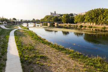 Fototapeta na wymiar Views at sunset of the French city of Beziers, with trees and one of its bridges reflected over the river Orb, and the 13th-century Cathedral of Saint Nazaire in the background