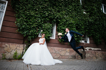 Elegant bride posing against the ivy wall while handsome groom jumping with a bouquet next to her.