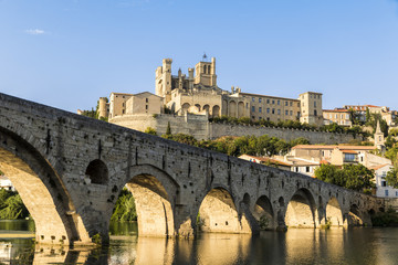 Fototapeta premium Views at sunset of the French city of Beziers, with trees and the old bridge reflected over the river Orb, and the 13th-century Cathedral of Saint Nazaire in the background