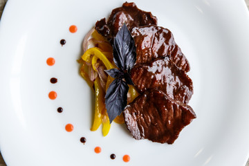 A white plate with slices of beef, stewed yellow pepper and onion. Purple basil on top. Drops of sauce.